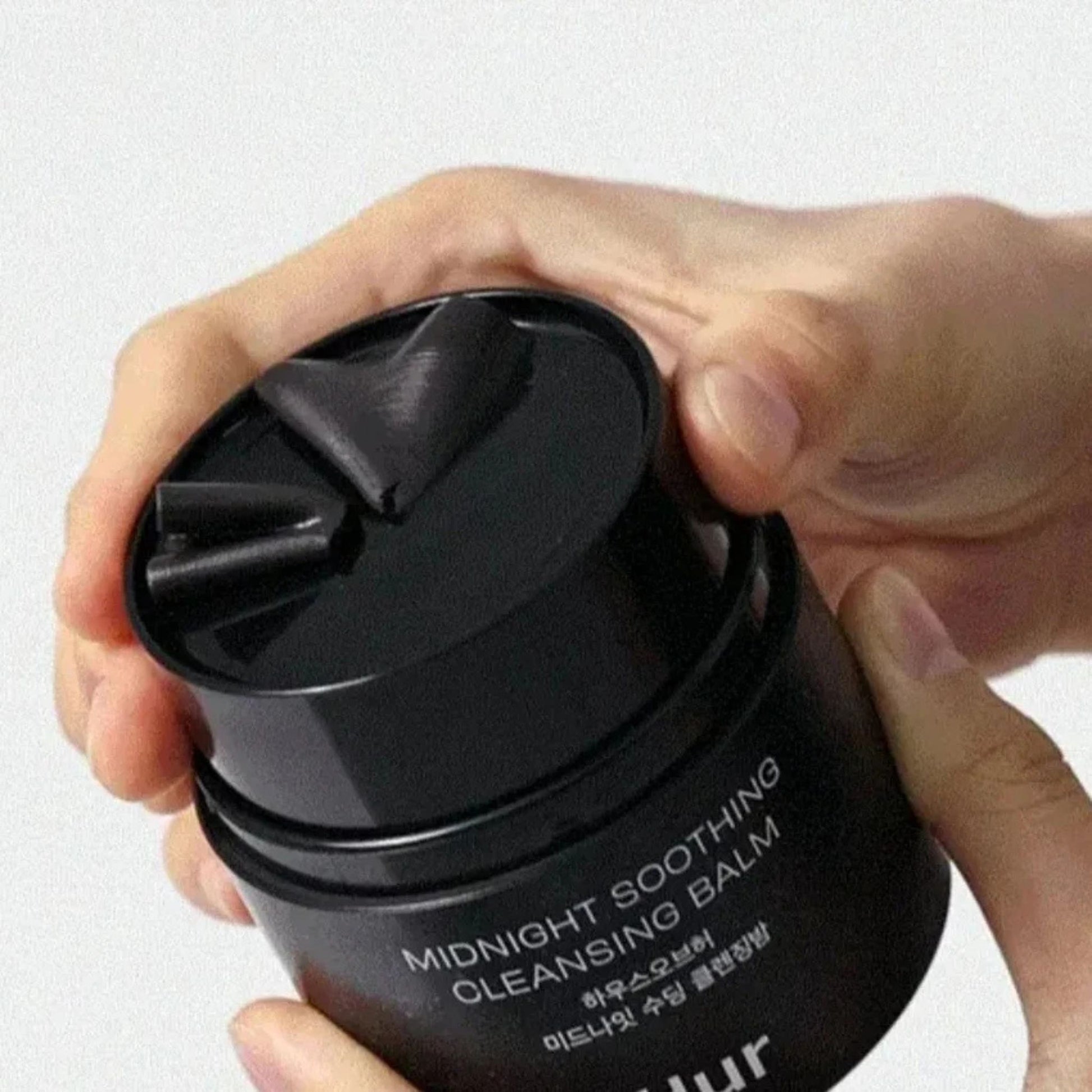 Black cleansing balm jar with 'Midnight Soothing Cleansing Balm' text held by a hand on a white background